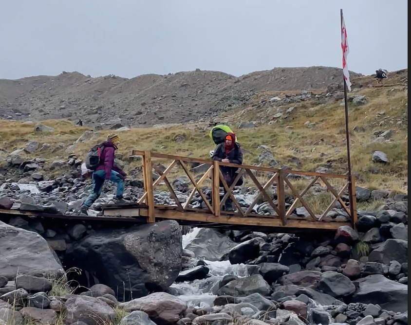 A woman crossing a bridge meets a man trekking to Mount Kazbek